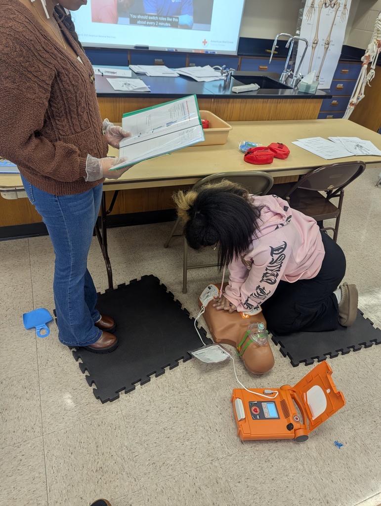 A student practices CPR on a training mannequin connected to an AED while a student stands beside her holding training materials. A Red Cross instructional video is displayed on a screen in the background.