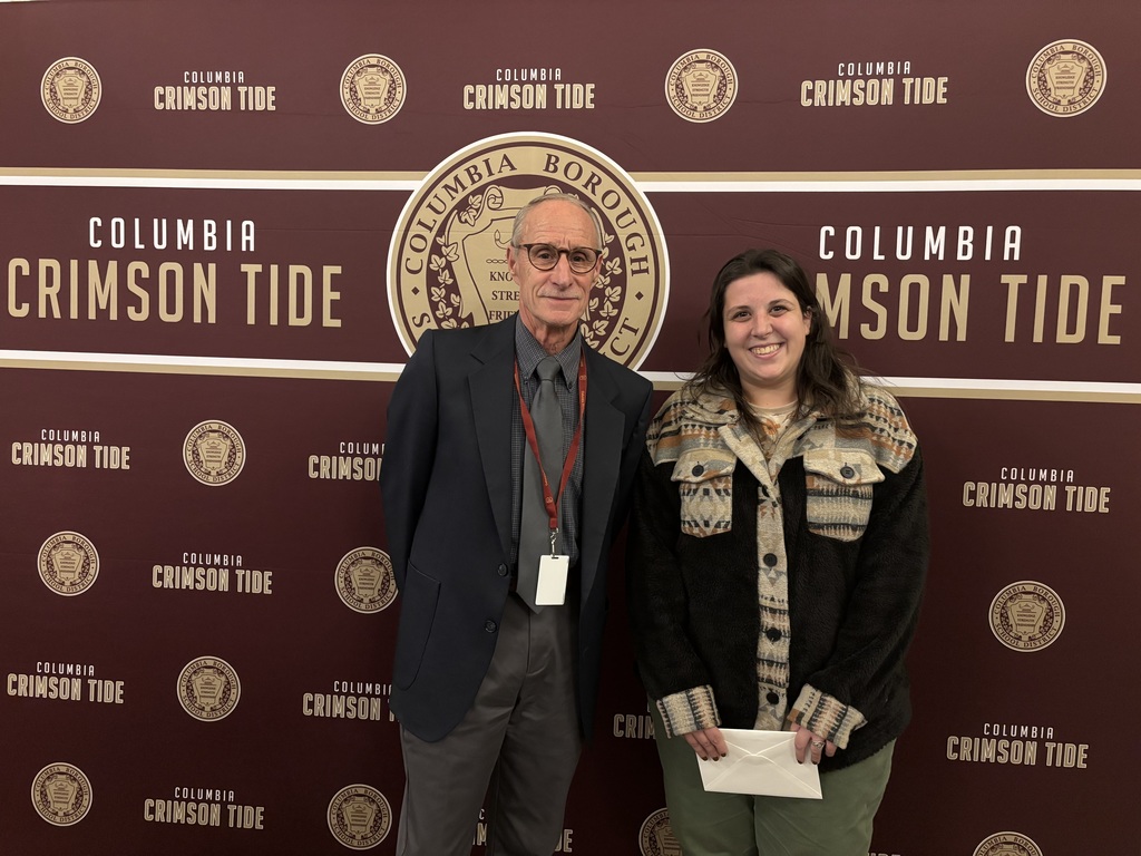 Two adults stand side by side in front of a maroon backdrop decorated with the Columbia Borough School District seal and the words ‘Columbia Crimson Tide.’ The person on the left, who appears to be a school board official, wears glasses, a dark suit jacket, and a lanyard. The person on the right, who appears to be a teacher, is smiling and wearing a patterned fleece jacket and green pants while holding a white envelope.