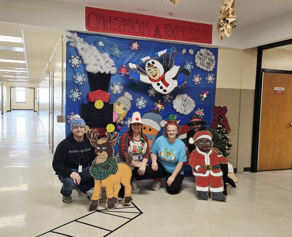 Four Hill Campus staff members kneel and smile in front of a large winter-themed backdrop outside the cafeteria. The display features snowflakes, a cartoon snowman, winter characters, and a Santa cutout that resembles the Hill Campus assistant principal. A small decorated Christmas tree stands nearby, and the hallway behind them extends into the school.