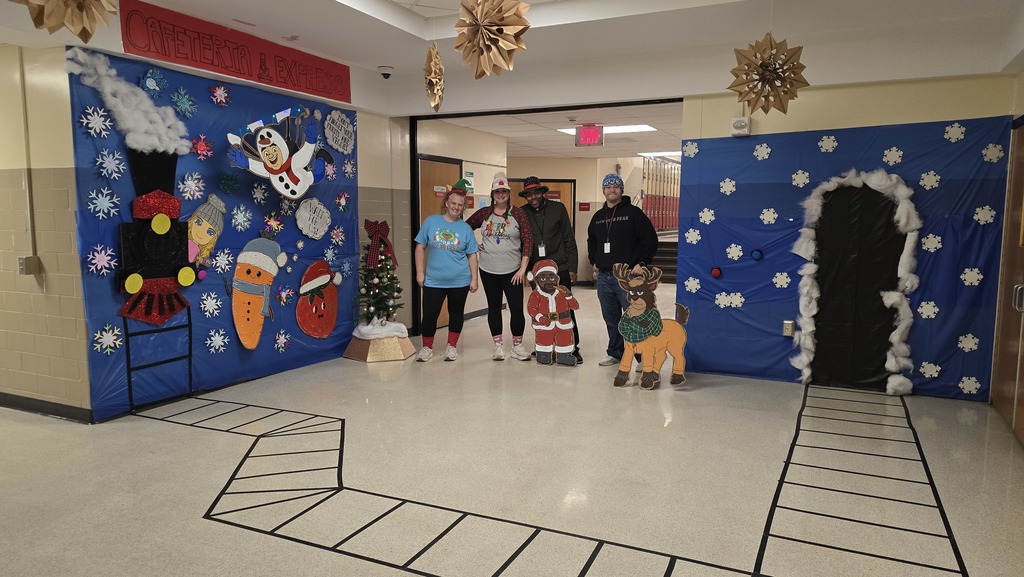 Four Hill Campus  staff members pose together in a school hallway between two winter-themed cafeteria displays. The decorations include blue backdrops with snowflakes, a Santa cutout, a reindeer cutout, and a festive winter scene. Paper snowflakes hang from the ceiling, and the taped train-track design continues along the floor, adding to the holiday theme.