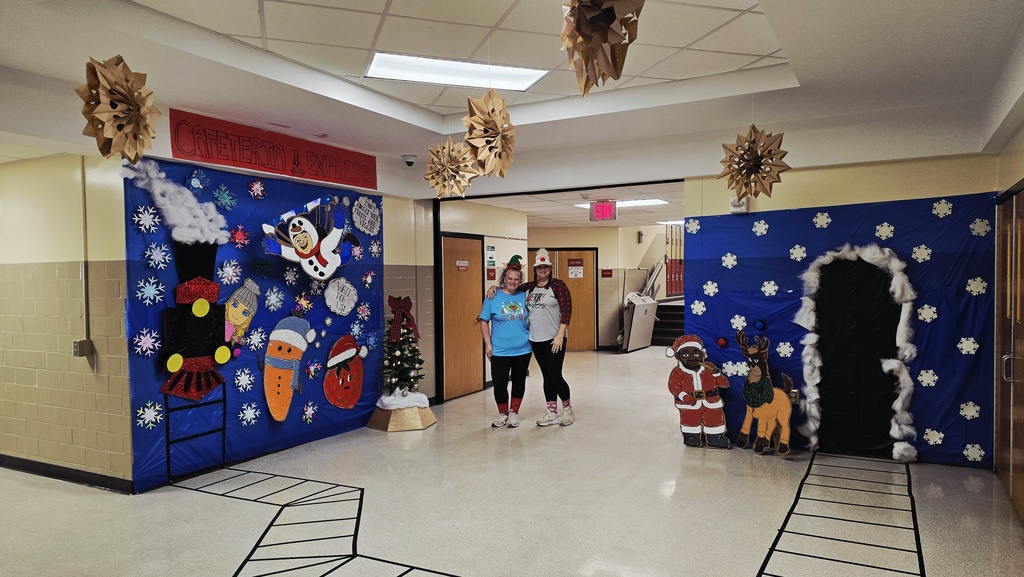 Two smiling Hill Campus cafeteria staff members stand in the center of a festively decorated hallway outside the cafeteria. On both sides are large blue winter-themed wall displays with snowflakes, cartoon characters, Santa, and a reindeer. Hanging paper snowflakes decorate the ceiling, and the taped train-track path is visible on the floor in front of them.