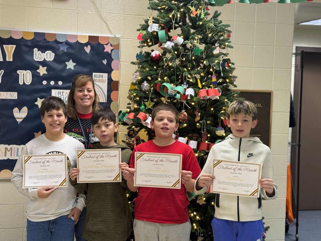 Five students stand in front of a decorated Christmas tree at Columbia Middle School – Taylor Campus, holding Student of the Month certificates, with a smiling staff member standing behind them. The students are dressed casually, and the certificates recognize them for December’s theme of empathy. The tree is decorated with lights, ornaments, and colorful paper chains, creating a festive school setting.