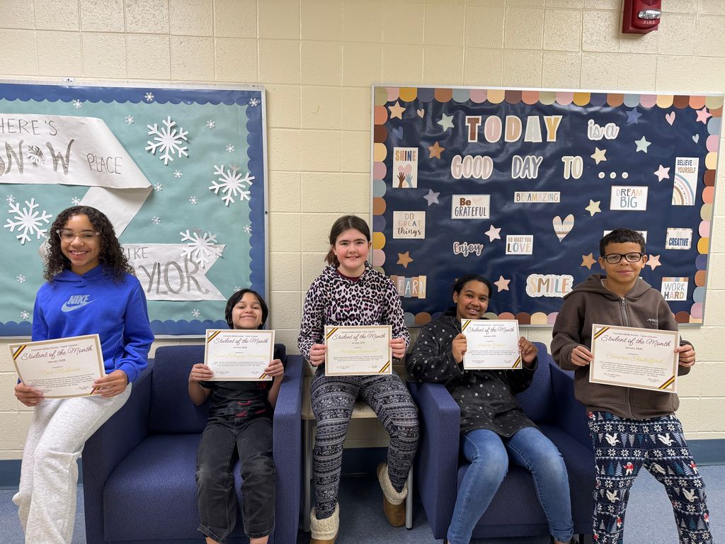 Four smiling children sit on blue chairs, proudly holding "Student of the Month" certificates. Behind them, colorful bulletin boards feature motivational messages and winter decorations.