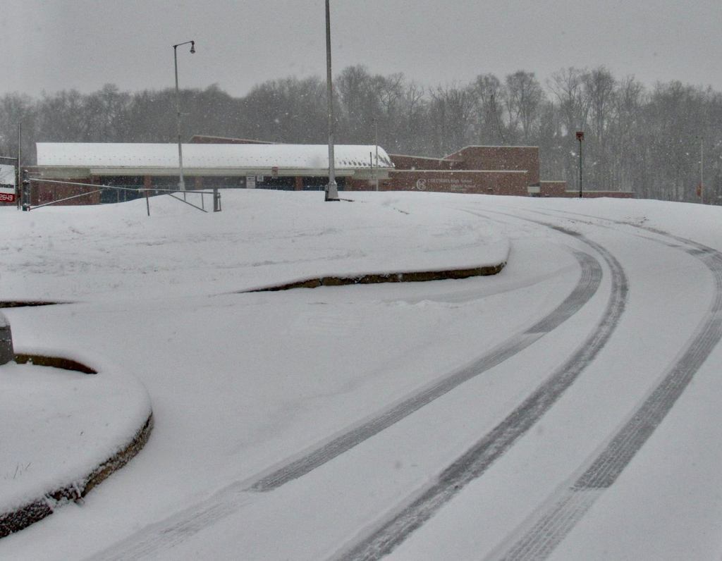 Snow-covered roadway on Columbia High School Middle School Hill Campus, showing curved tire tracks in the foreground, empty sidewalks and parking areas, and school buildings in the background as light snow falls under an overcast sky.