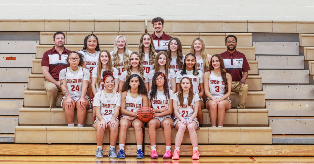 Columbia High School Crimson Tide girls basketball team poses for a formal team photo inside a gymnasium. Fourteen players in white “Crimson Tide” uniforms with maroon lettering and numbers are arranged in three rows on wooden bleachers, with two coaches in maroon and gray polo shirts positioned at the ends and one coach standing centered behind the top row. The front row of players is seated, the middle row kneels or sits, and the back row stands. A basketball rests on the lap of a seated player in the center of the front row. The gym floor is polished wood, and the background shows light-colored cinderblock walls and folded bleachers. The team colors are maroon and gold.