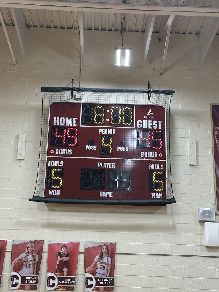 A gymnasium scoreboard displays the final score of a girls basketball game. The home team has 49 points and the guest team has 45 points. The scoreboard shows Period 4, team fouls at 5 for each side, and bonus indicators lit for both teams. The scoreboard is mounted on a light-colored cinderblock wall and covered with protective netting. Below the scoreboard, banners feature individual player posters with names and photos, including Brynn Bigler (#15), Mekhi Johnson-Myer, and Delaney Burke (#20). Overhead lights and exposed ceiling beams are visible above the scoreboard.