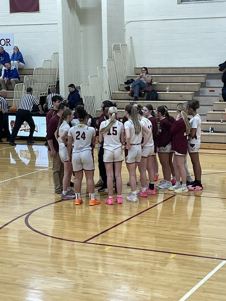 Columbia High School Crimson Tide girls basketball players stand in a tight huddle with their coaches on the gym floor during a game. The players wear white uniforms with maroon numbers and trim, along with various brightly colored basketball shoes. The coaches, dressed in maroon tops and khaki pants, stand within the circle speaking to the team. The players face inward, listening attentively with focused body language. In the background, referees in black-and-white striped shirts stand near the scorer’s table, and a few spectators sit on wooden bleachers against a light-colored gym wall.