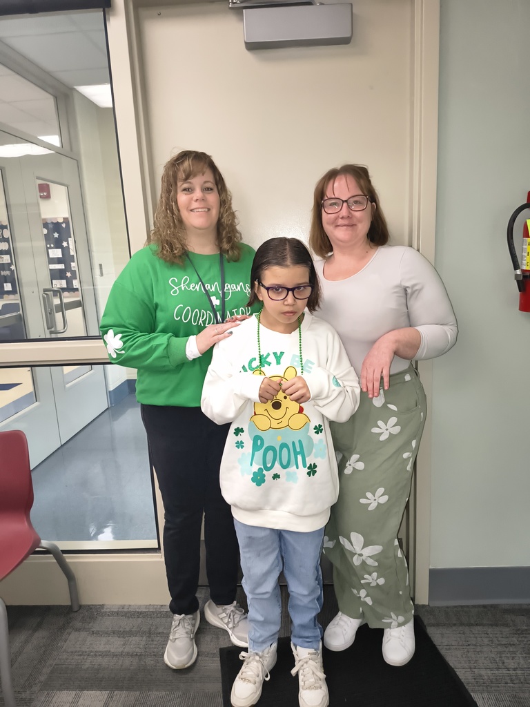 Three people stand together in a school hallway. Two adult women stand behind a young student. One woman wears a green shirt, and the other wears a light gray top with green patterned pants. The student in front wears glasses and a white Winnie the Pooh sweatshirt with green beads. All three are smiling or posing for the photo.