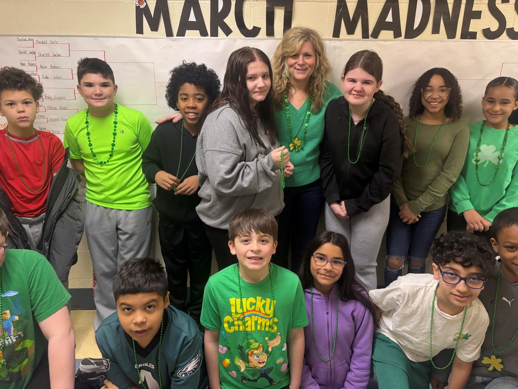 A group of middle school students and one adult pose together in front of a “March Madness” bracket display on a wall. Many students are wearing green clothing and green bead necklaces, suggesting a St. Patrick’s Day theme. The group is arranged in two rows, with some students standing and others crouching in front, all facing the camera.