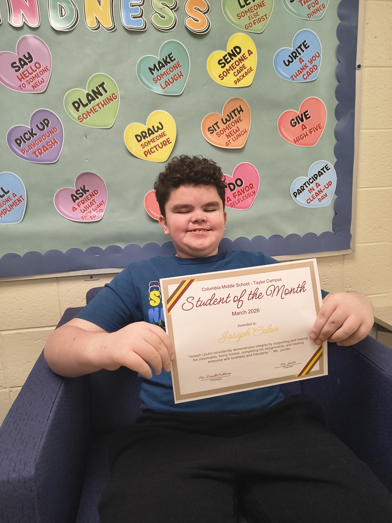 A Columbia Middle School – Taylor Campus student sits in a chair smiling while holding a “Student of the Month – March 2026” certificate with the name Joseph Colon. Behind him is a “Random Acts of Kindness” bulletin board decorated with colorful heart-shaped messages promoting kind actions.