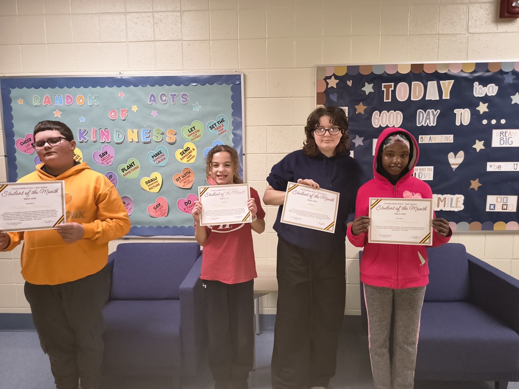 Four Columbia Middle School – Taylor Campus students stand side by side holding “Student of the Month – March 2026” certificates. They are posing in front of colorful bulletin boards, including one that reads “Random Acts of Kindness” with heart-shaped notes and another with positive messages. The students are smiling and dressed casually, celebrating their recognition for demonstrating integrity.