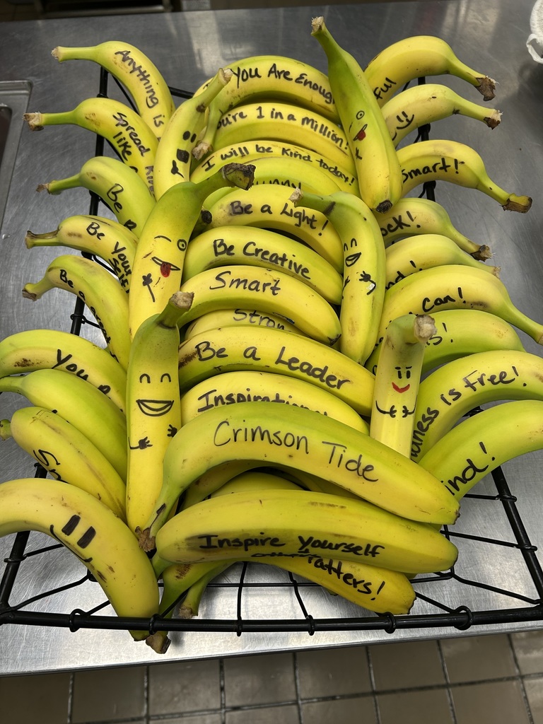 A basket of bananas on a countertop, each banana decorated with handwritten positive messages and simple smiley faces. Messages include phrases such as “You are enough,” “Be a leader,” “Be creative,” “Be a light,” “Smart,” “Strong,” and “Crimson Tide.” The display appears to promote encouragement and positivity.