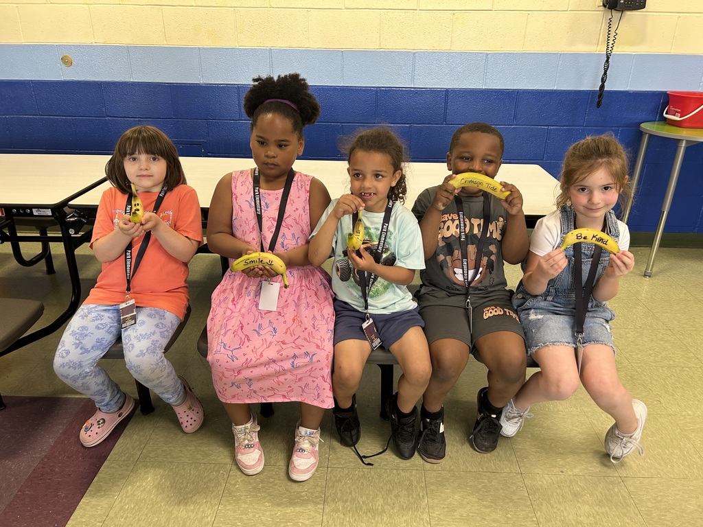 Five young children sit side by side on a cafeteria bench, each holding a banana decorated with handwritten positive messages. The children smile and display messages such as “Smile,” “Crimson Tide,” and “Be Kind,” highlighting a fun activity focused on encouragement and positivity.