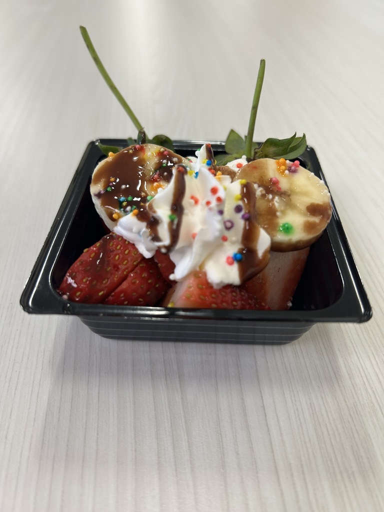 A small black tray filled with sliced bananas and strawberries topped with whipped cream, chocolate drizzle, and colorful sprinkles. Two strawberry stems extend upward, and the dish is arranged as a decorative fruit dessert.