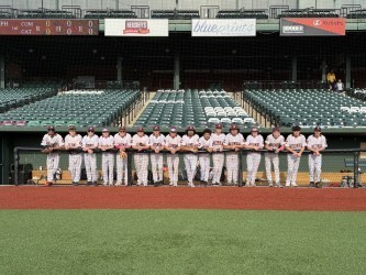 A baseball team in white uniforms stands in a straight line across the field in front of empty stadium seats, posing together for a group photo during daylight.