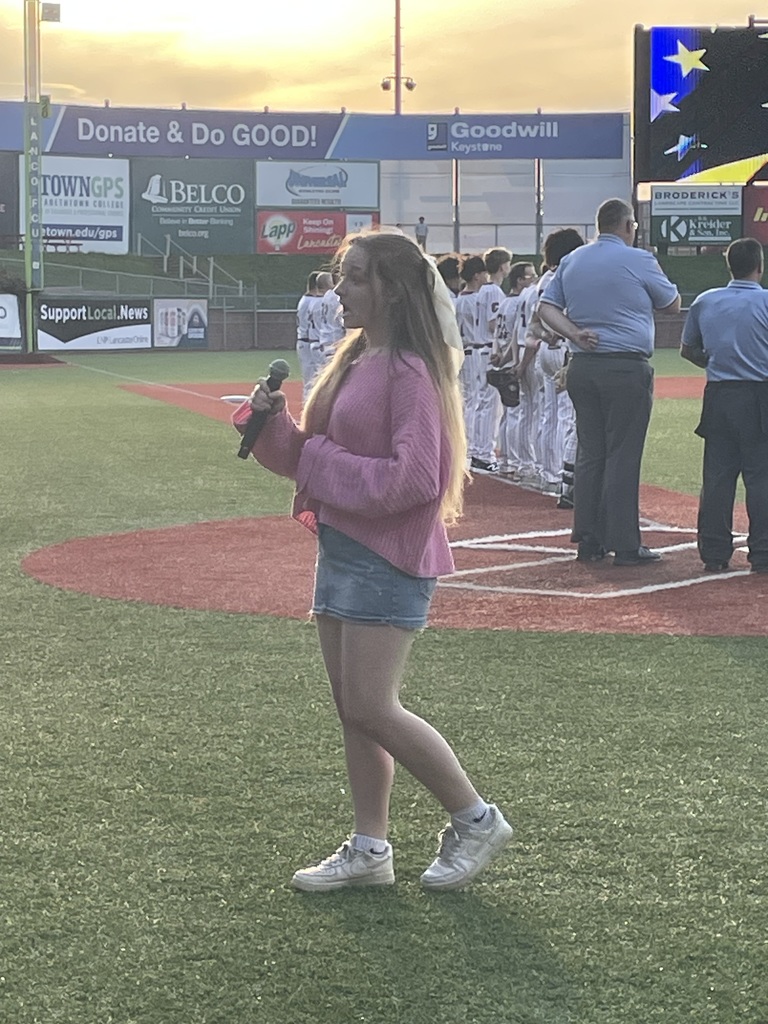 A young woman stands on a baseball field holding a microphone, singing during a pregame ceremony. She wears a pink top, denim shorts, and sneakers, with a team lined up in the background near the baseline.