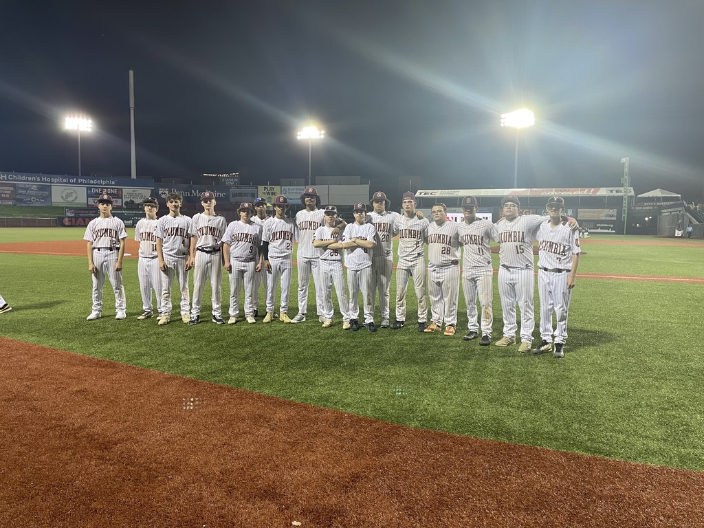 A baseball team in white uniforms stands in a line on the field at night under bright stadium lights, facing forward as if being introduced before a game.