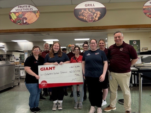 A group of school staff members stand together in a cafeteria kitchen holding a large ceremonial check from GIANT. The check is made out to Columbia School District for $2,067.95 and dated April 16, 2024. Overhead signs read “Fruits & Vegetables” and “Grill.” The group smiles at the camera, indicating a donation or partnership event.