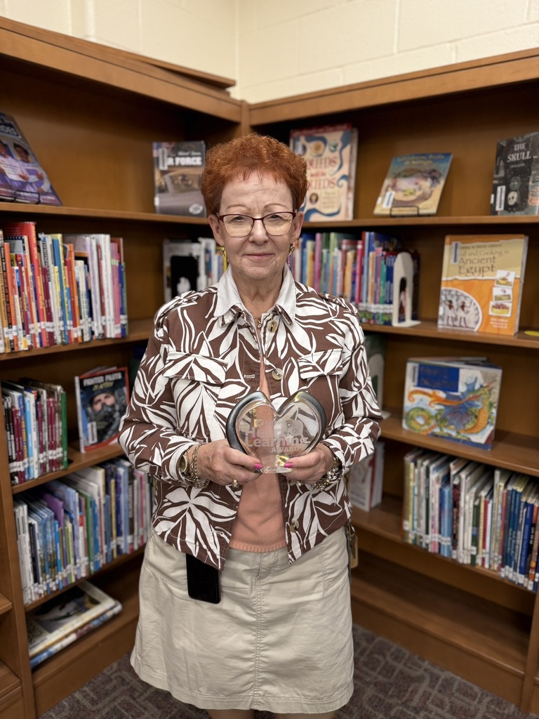 Arlene Gibble stands  in a school library holding a heart-shaped award. She smiles at the camera, standing in front of bookshelves filled with children’s books, representing recognition for an educational achievement or award.