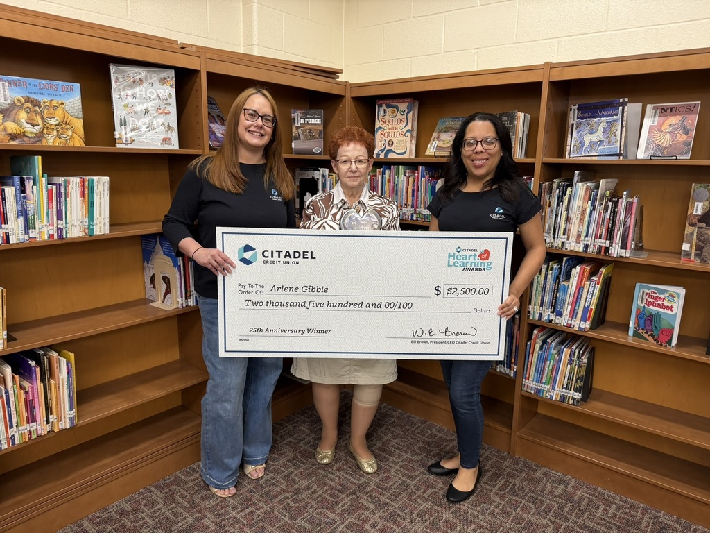 Three women stand in a school library holding a large ceremonial check from Citadel Credit Union. The check is made out to Arlene Gibble for $2,500 as a Heart of Learning Award winner. Bookshelves filled with children’s books line the background.