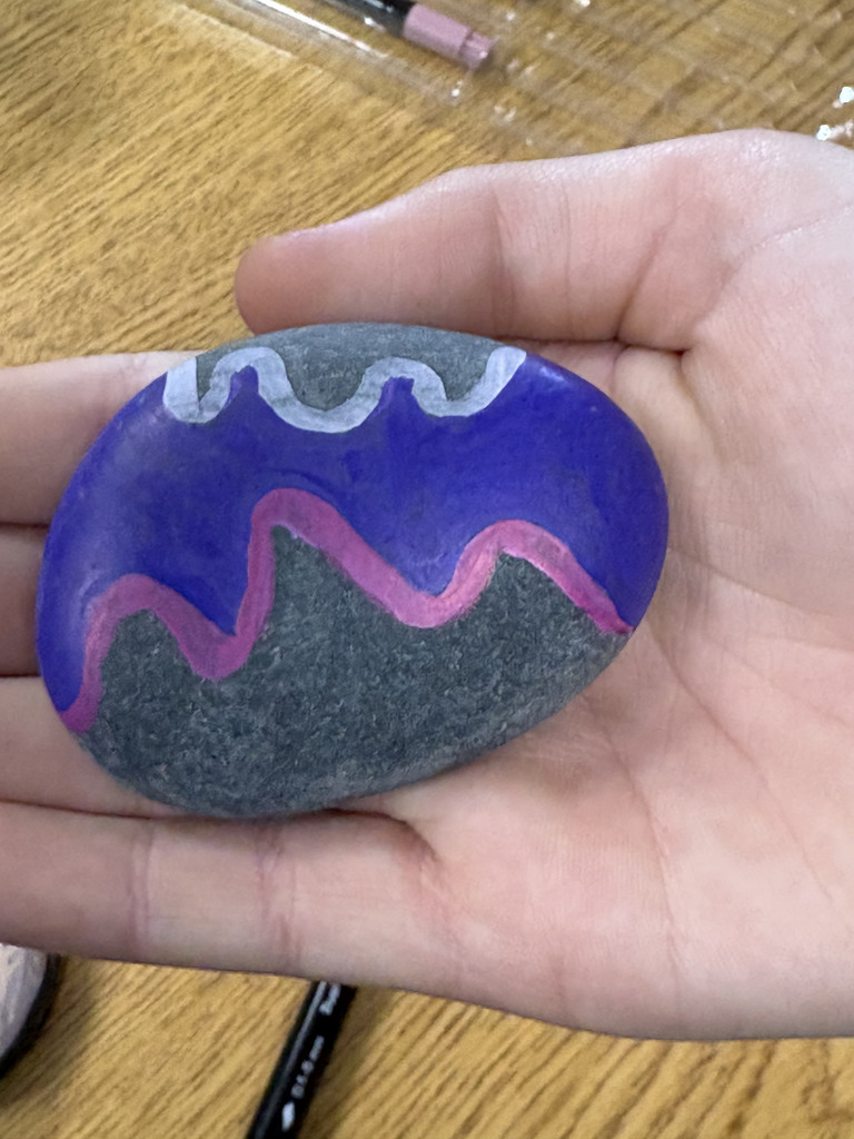 A hand holds a smooth rock painted with bold purple and pink wavy designs across the top.