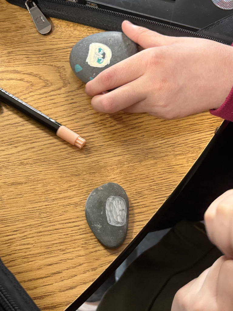 Close-up of a student’s hand holding a painted rock with a small design, while another painted rock rests on the desk beside a marker.
