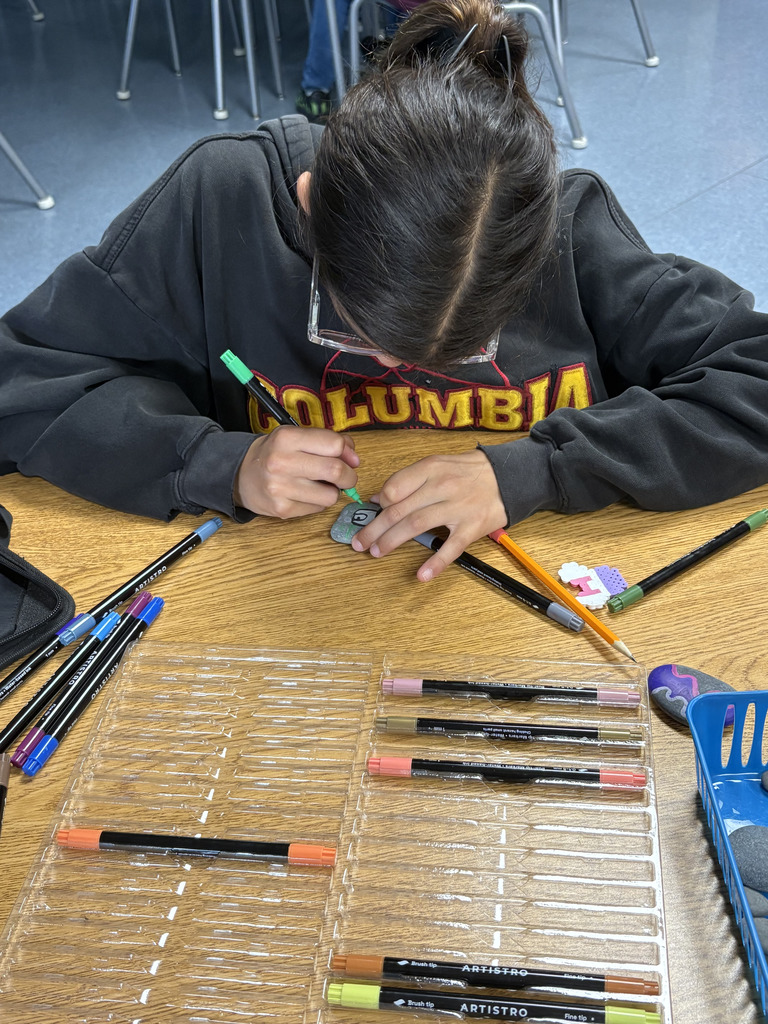 Student wearing a “Columbia” sweatshirt leans over a desk, carefully drawing on a small rock with a green marker. Art supplies, including markers and pencils, are spread across the table.