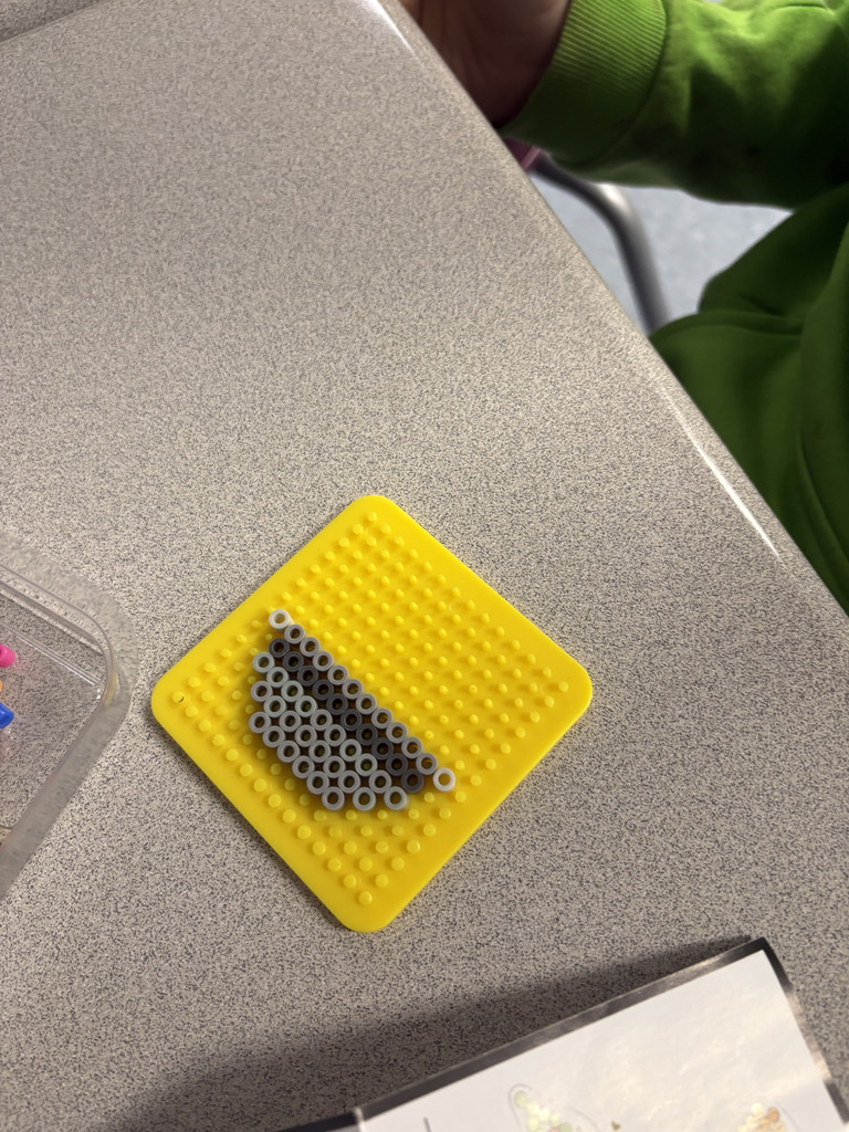 A yellow pegboard with small gray beads arranged in a triangular pattern sits on a classroom desk. A student in a green sleeve is partially visible nearby.