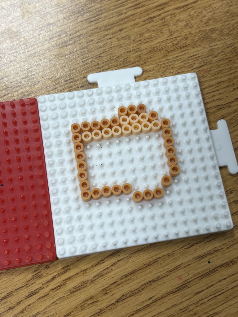 A pegboard with tan beads arranged in a square outline with a small raised section at the top sits on a desk next to another pegboard.