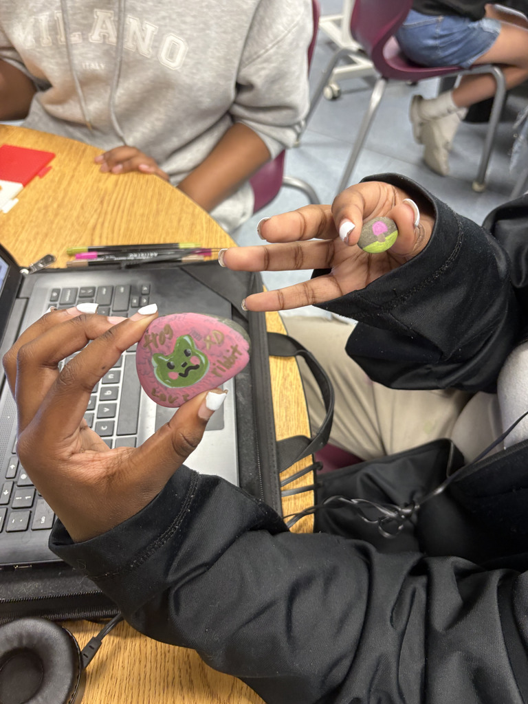 Two students hold up decorated rocks. One rock features a green cartoon-style face with writing around it, while the other shows a small, colorful design. A laptop and markers are on the desk.