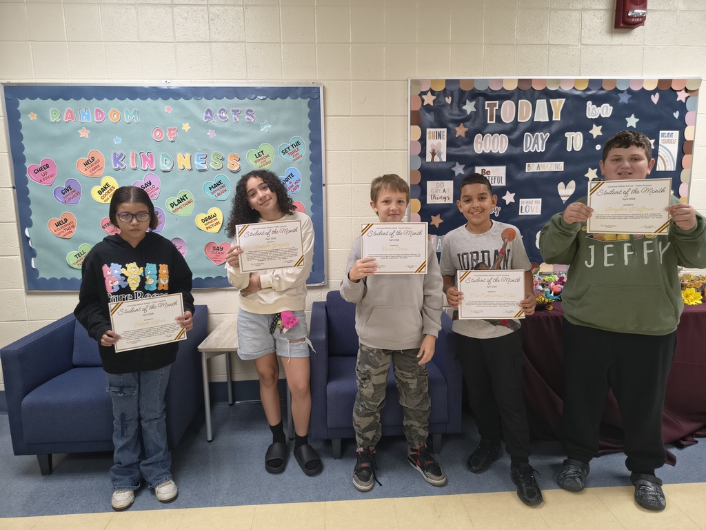 Five students stand side by side indoors holding “Student of the Month” certificates. They are smiling and posing in front of two colorful bulletin boards—one reads “Random Acts of Kindness” with heart-shaped messages, and the other says “Today is a Good Day To…” with positive phrases. The students are dressed casually, and a small table with decorations is visible to the right.