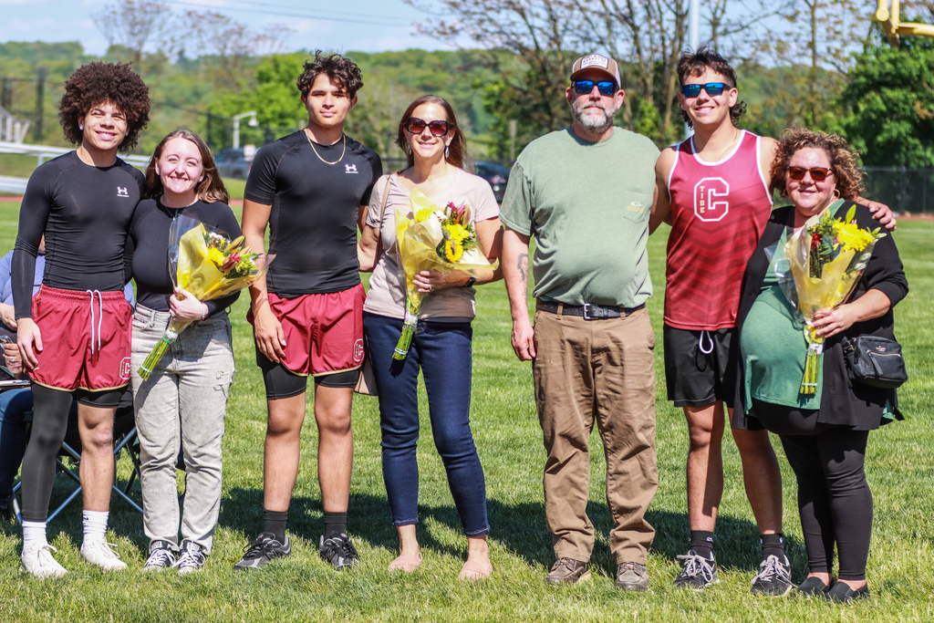 Group photo of track and field senior athletes standing on a grassy field with family members, smiling and holding bouquets of flowers during a senior recognition event outdoors on a sunny day.