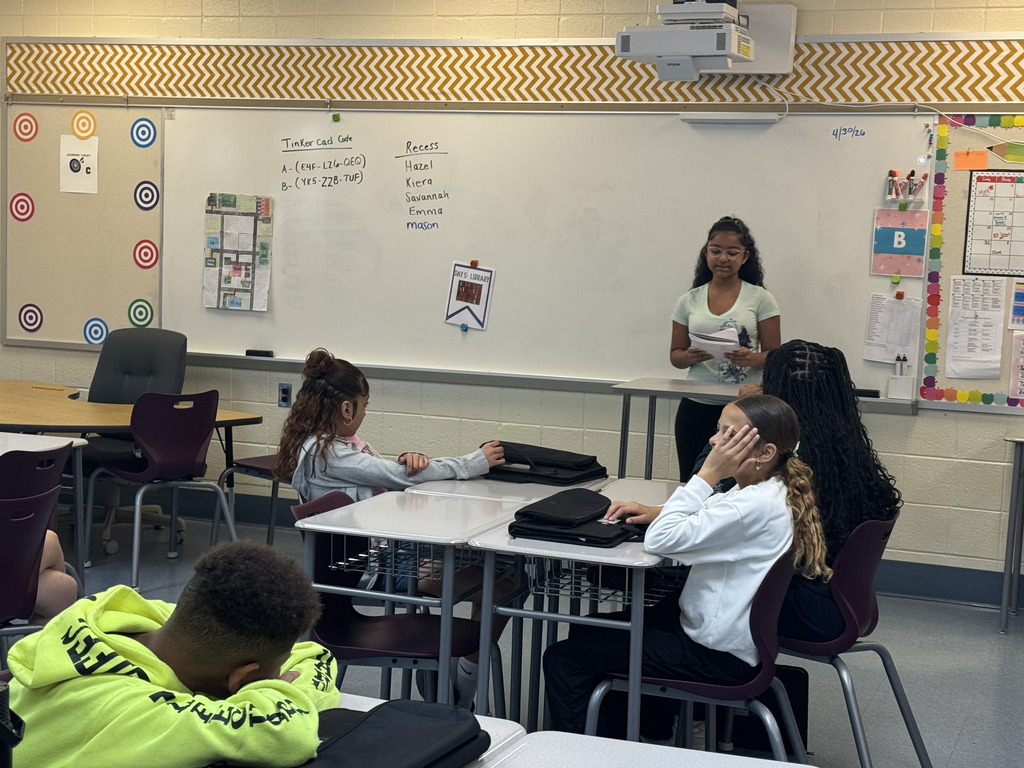 A student stands at the front of a classroom reading from a paper while four other students sit at desks listening. A whiteboard with notes is visible behind her.