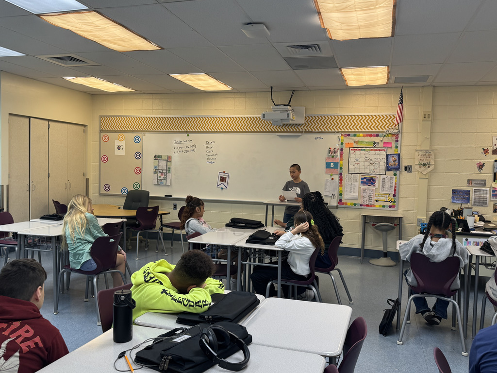 A student stands at the front of a classroom giving a presentation while classmates sit at tables listening or resting their heads.