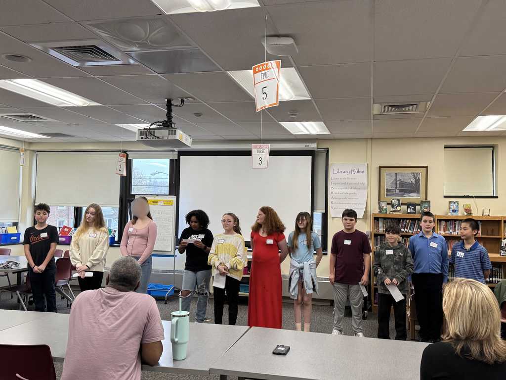 A group of middle school students stand at the front of a school library or classroom during a presentation or event. The students are lined up in a row holding papers, while adults sit at tables watching. Hanging number signs are suspended from the ceiling above them. Bookshelves, windows, and a projector screen are visible in the background. One student’s face is blurred for privacy.