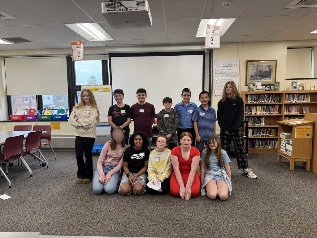 A group of middle school students stand at the front of a school library or classroom during a presentation or event. The students are lined up in a row holding papers, while adults sit at tables watching. Hanging number signs are suspended from the ceiling above them. Bookshelves, windows, and a projector screen are visible in the background. One student’s face is blurred for privacy.