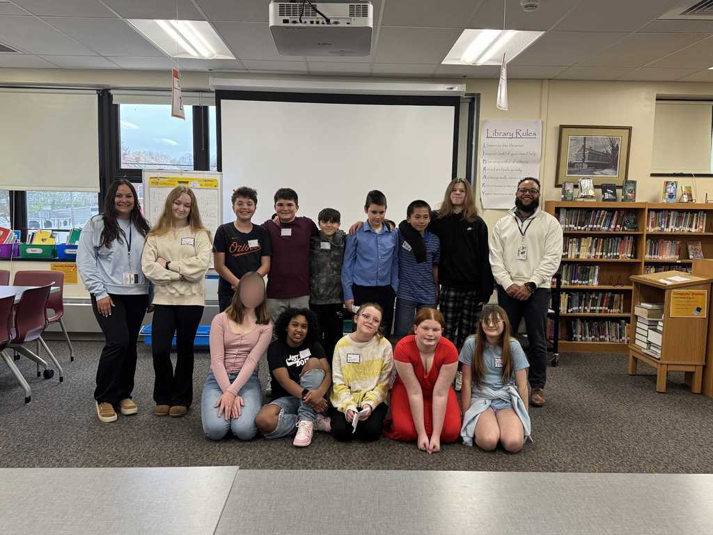 A group of middle school students stand at the front of a school library or classroom during a presentation or event. The students are lined up in a row holding papers, while adults sit at tables watching. Hanging number signs are suspended from the ceiling above them. Bookshelves, windows, and a projector screen are visible in the background. One student’s face is blurred for privacy.