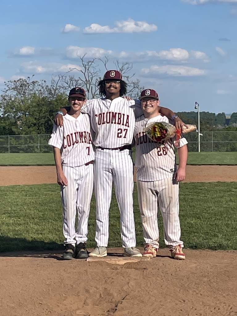 Three Columbia baseball seniors standing together on the pitcher’s mound during Senior Night. The players are wearing white pinstriped Columbia uniforms and maroon caps, smiling with their arms around each other. One player is holding a bouquet of flowers. A baseball field, green grass, and blue sky with scattered clouds are visible in the background.