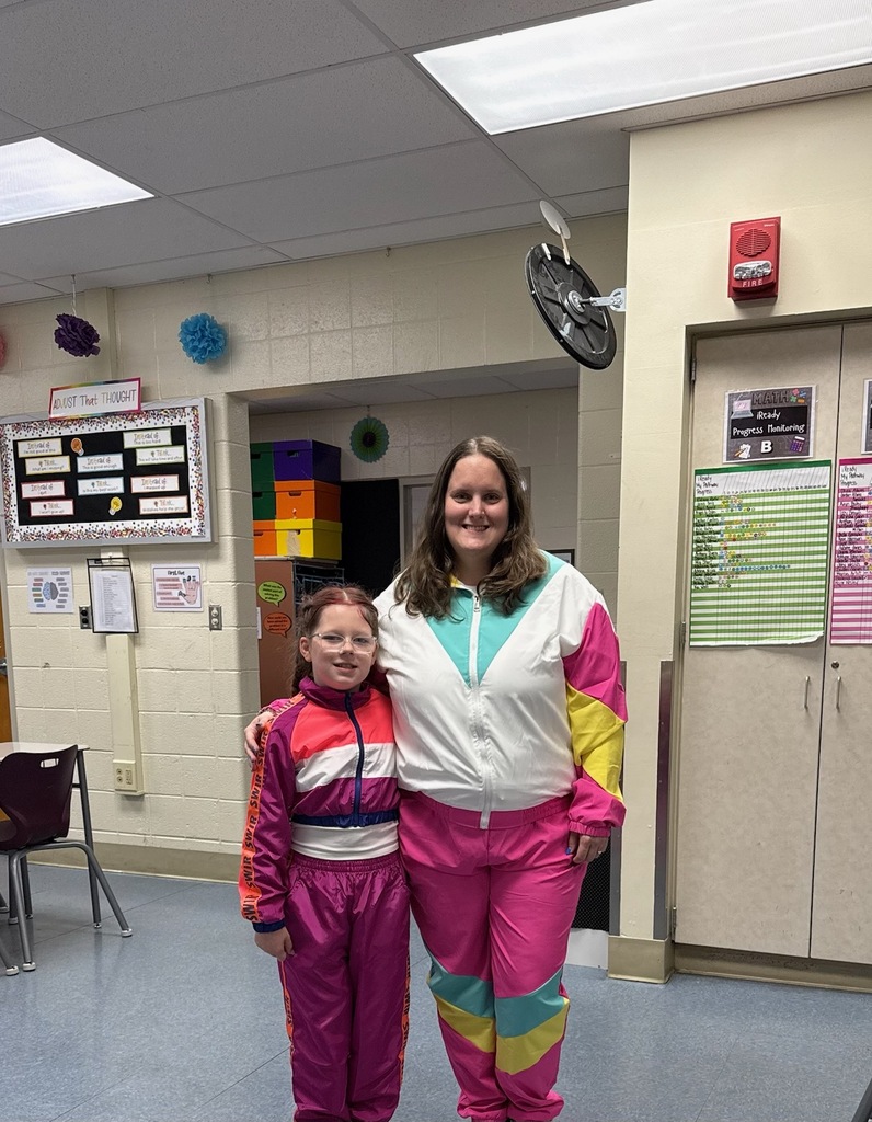 Three people standing together indoors, smiling at the camera in a classroom or school setting. The person on the left is wearing denim short overalls over a pink shirt with a large blue bow in their hair and holding up a peace sign. The person in the middle is wearing a colorful retro-style tracksuit with pink, teal, yellow, and white accents. The person on the right is wearing sunglasses, a white-and-pink tie-dye shirt, ripped denim shorts, and black shoes. Tables, chairs, and classroom supplies are visible in the background.