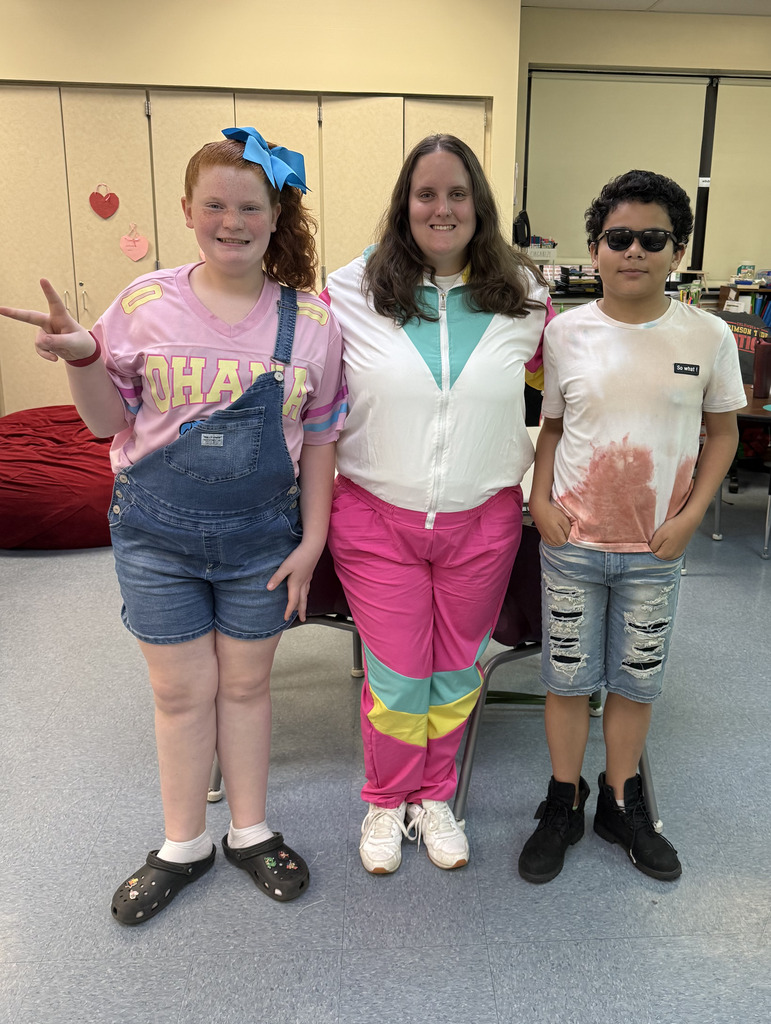 Three people standing together indoors, smiling at the camera in a classroom or school setting. The person on the left is wearing denim short overalls over a pink shirt with a large blue bow in their hair and holding up a peace sign. The person in the middle is wearing a colorful retro-style tracksuit with pink, teal, yellow, and white accents. The person on the right is wearing sunglasses, a white-and-pink tie-dye shirt, ripped denim shorts, and black shoes. Tables, chairs, and classroom supplies are visible in the background.