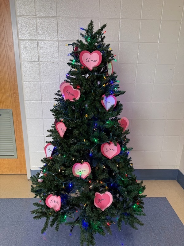 A Christmas tree decorated with pink heart-shaped cutouts, each bearing a name. The tree is located in an indoor space with walls and flooring visible.