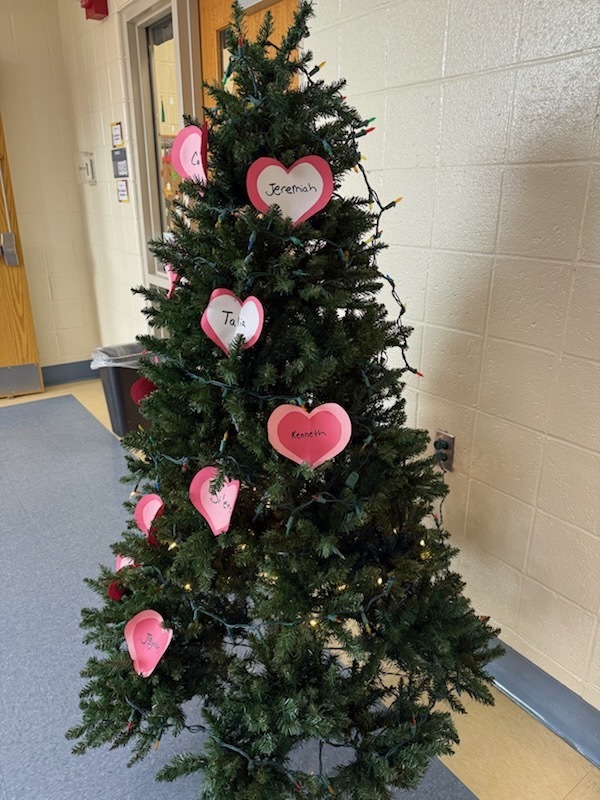 A Christmas tree decorated with pink heart-shaped cutouts, each bearing a name. The tree is located in an indoor space with walls and flooring visible.