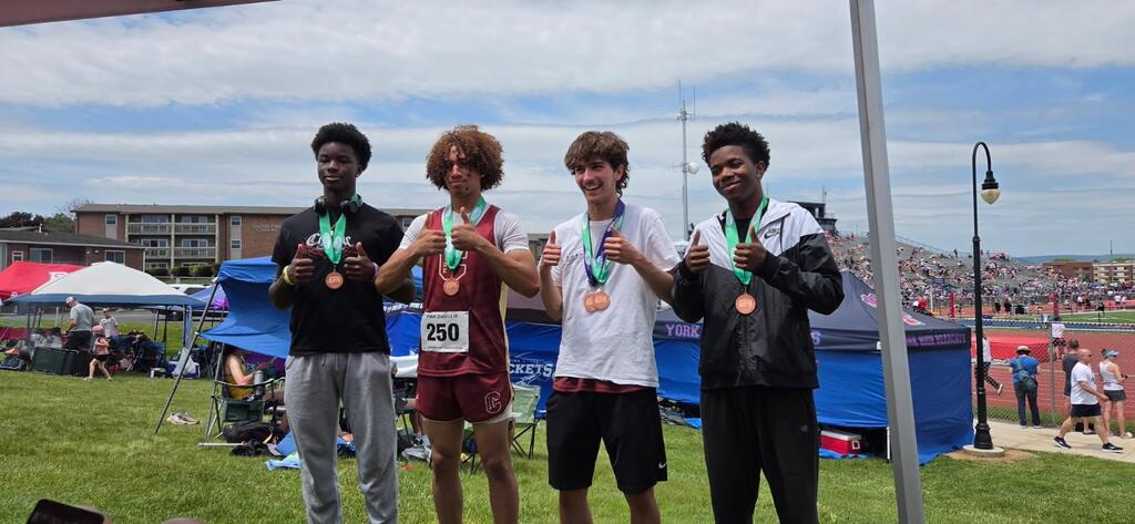 Four high school track athletes wearing medals stand side by side on a grassy area near the track at the District 3 Track & Field Meet. Each athlete is smiling and giving a thumbs-up gesture. Tents, spectators, and the track stadium are visible in the background under a partly cloudy sky.