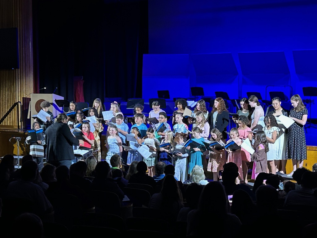 A children's choir performs on stage with a conductor, against a blue-lit backdrop.