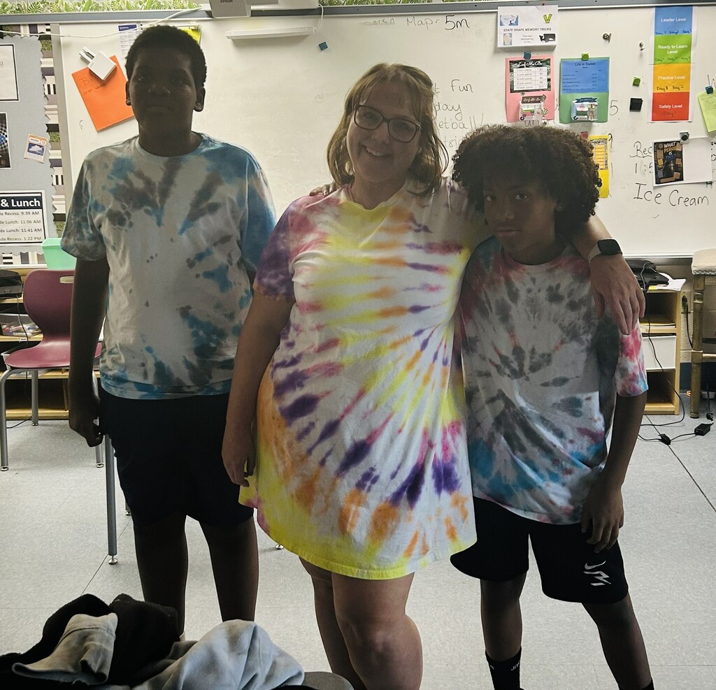Three people stand together in a classroom, each wearing colorful tie-dye shirts. They smile warmly, conveying a sense of unity and joy.