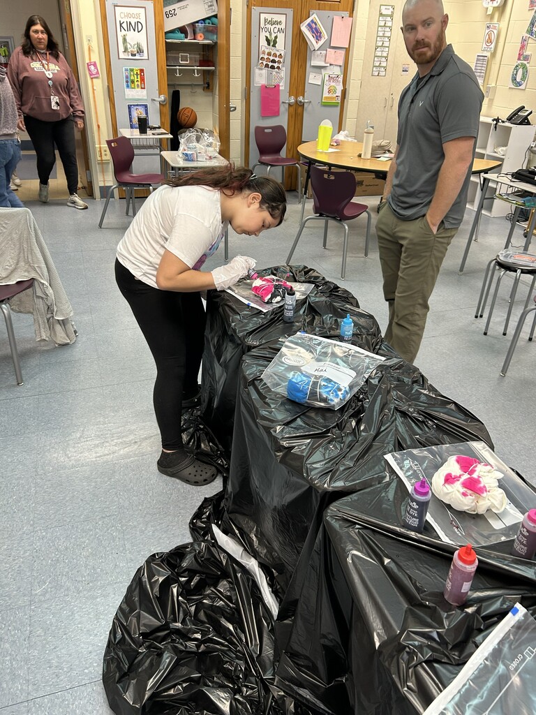 A girl, wearing gloves, dyes fabric with colorful liquids on plastic-covered tables in a classroom. A man observes. Bright, creative, energetic scene.