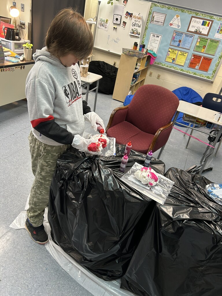A child in a gray hoodie and camouflage pants is tie-dyeing shirts in a classroom. Black plastic covers the table, and colorful dye bottles are scattered around.