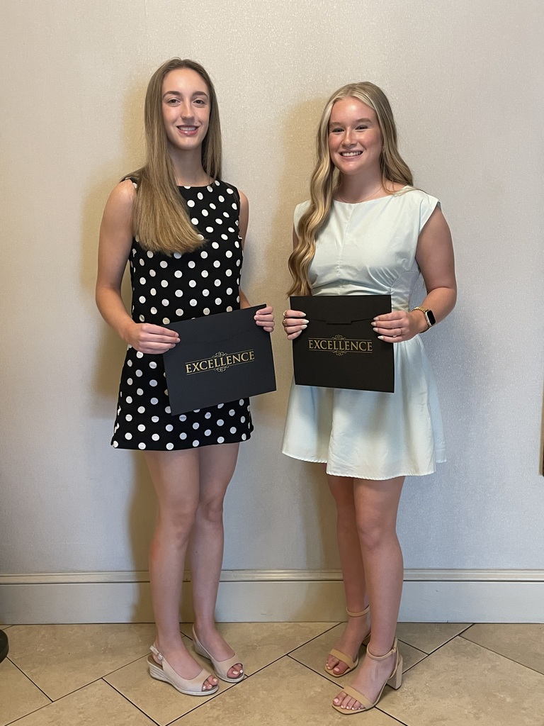 Two young women stand side by side indoors holding black folders with the word “EXCELLENCE” in gold lettering. The student on the left is wearing a black sleeveless dress with white polka dots and beige wedge sandals. The student on the right is wearing a light blue sleeveless dress with tan heeled sandals and a smartwatch. Both are smiling while posing at the A. Landis Brackbill Scholar Athlete Award ceremony.