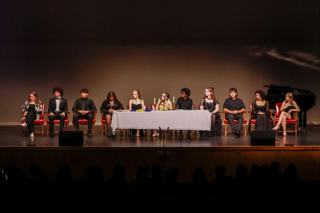 Students dressed in formal black attire sit across a stage during a theater awards and induction ceremony. A long table with candles is centered onstage, with audience silhouettes visible in the foreground.