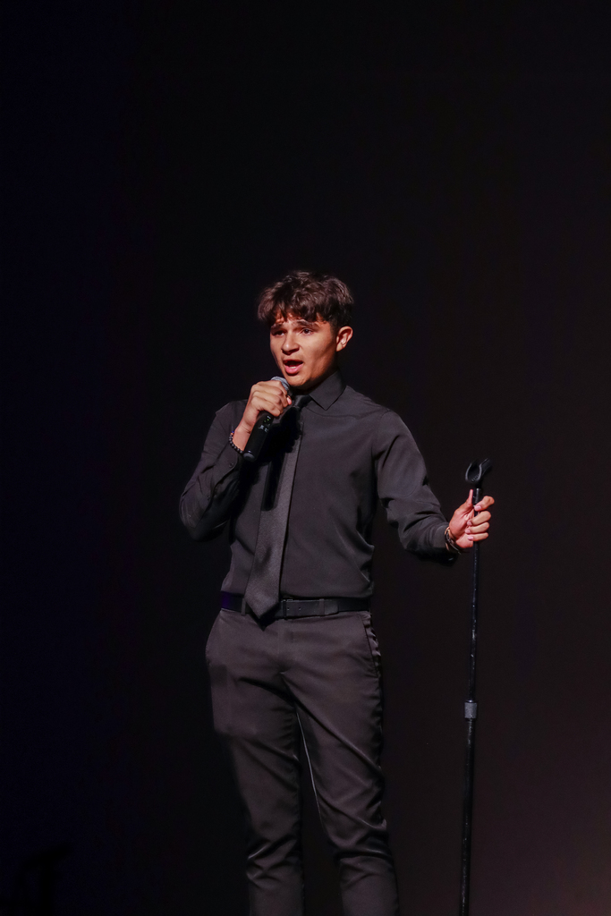 A student performer dressed in black formal attire sings into a microphone on a darkened stage during a theater showcase performance.